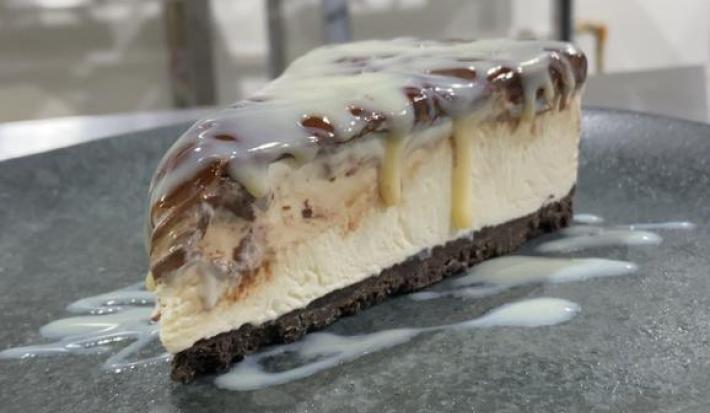 Foto da receita de CHEESECAKE MOÇA GANACHE. Observa-se uma fatia da cheesecake em um prato de cerâmica preto, com Leite MOÇA escorrendo pelo recheio.