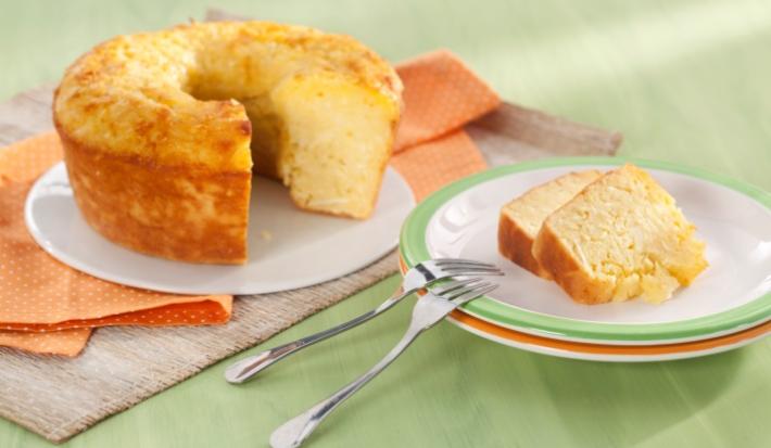 Fotografia em tons de laranja e verde em uma mesa de madeira verde com um pano laranja de bolinhas branca. Ao centro, um prato raso branco com o bolo de macaxeira cortado. Ao lado, um prato branco com borda verde e duas fatias do bolo.