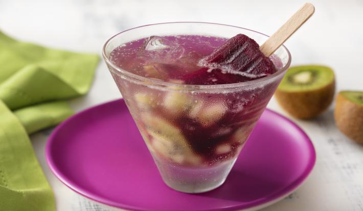 Fotografia em tons de branco, roxo e verde de uma bancada de madeira branca, um paninho verde e um prato redondo roxo, sobre ele um copo redondo de vidro com o drink de picolé. Ao fundo um kiwi cortado ao meio.