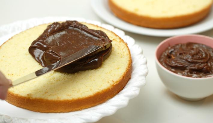 Foto de um bolo aberto ao meio em cima de um prato branco sendo preenchido com creme de chocolate por cima Foto de um bolo aberto ao meio em cima de um prato branco sendo preenchido com creme de chocolate por cima