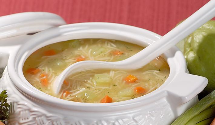 Fotografia em tons de branco, vermelho e verde, com sopeira branca ao centro com sopa de legumes, macarrão e um concha branca, tendo em volta chuchu, cebolinha, cenoura, cebola, salsa e uma tampa branca, tudo sobre bancada vermelha.