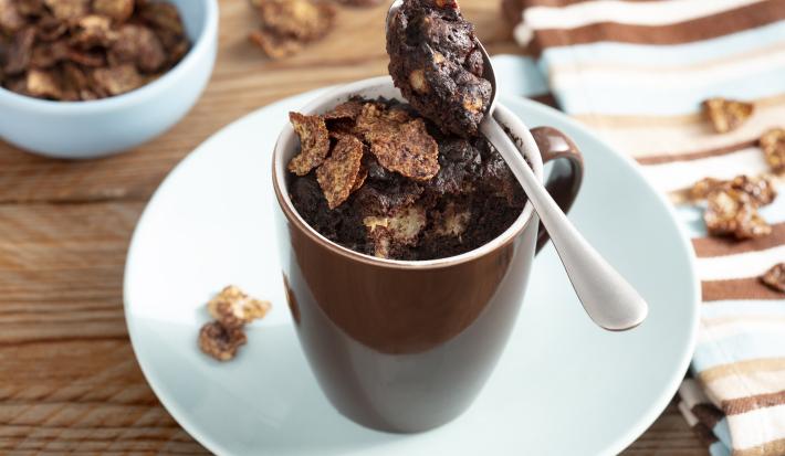Fotografia em tons de marrom em uma bancada de madeira com um pano listrado em marrom, azul claro e bege ao lado e ao centro, um prato branco com uma xícara marrom em cima dele e o bolo de caneca de chocolate dentro, decorado com os cereais Nesfit.