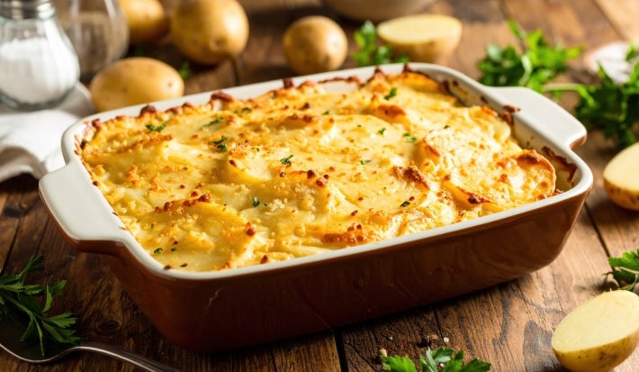 Em cima de uma mesa, refratário retangular com batatas e queijo gratinado, decoado com cheiro-verde picado.