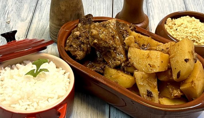 Foto em tons de marrom da receita de costela na pressão servida com batatas couradas em uma travessa com material que imita barro. Ao lado, uma panelinha vermelha com arroz branco e um recipiente com farofa