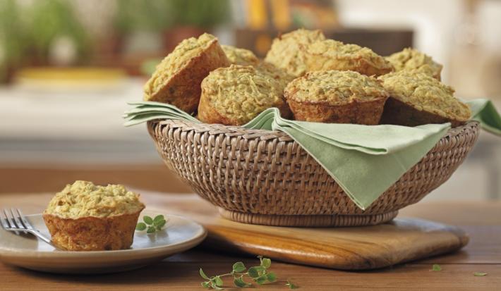 Fotografia em tons de verde em uma bancada de madeira, uma tábua de madeira, uma cesta de vime com vários muffins de aveia e embaixo um paninho verde. Ao lado, um pratinho de sobremesa branco com um muffin em cima dele.