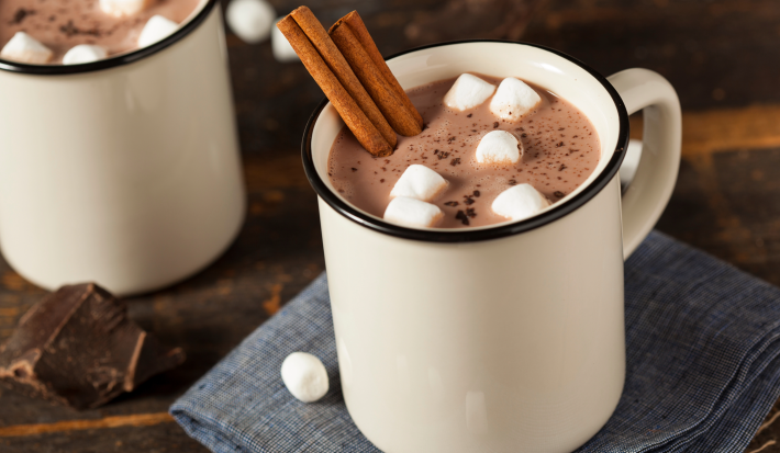 Imagem de uma caneca branca com chocolate quente, pedaços de marshmallow e 2 pedaços de canela em pau.