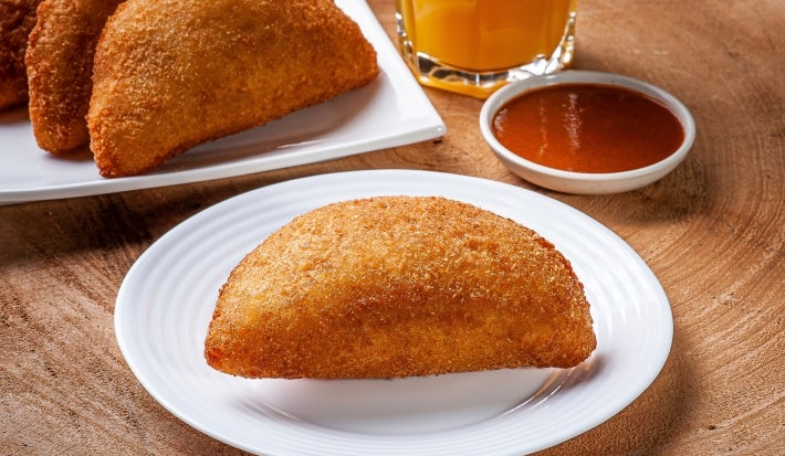 Fotografia de um risole de presunto e queijo em um prato raso branco sobre a mesa de madeira. Ao fundo tem suco de laranja, o molho e mais risoles.