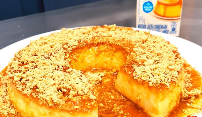 Fotografia de um pudim de paçoca com caramelo e paçoca triturada por cima. O pudim está sobre um prato grande e raso branco.
