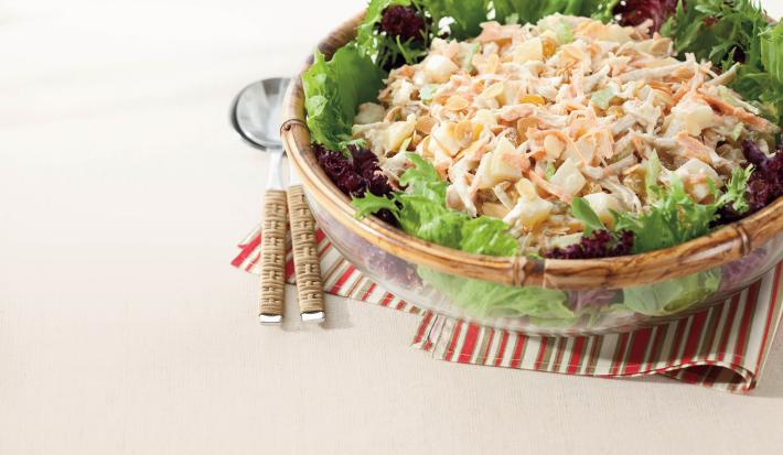 Fotografia em tons de rosa e bege, com saladeira ao centro com folhas verde e roxas e salada de frango cremosa sobre guardanapo com listras coloridas, duas colheres de servir ao lado, tudo sobre toalha rosa.