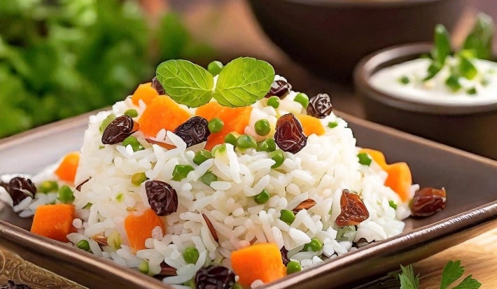 Em um prato de porcelana marrom, há uma porção de arroz branco misturado com cenouras, ervilhas e uvas-passas, decorado com duas folhas de hortelã.