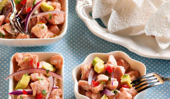 Fotografia em tons de azul em uma bancada com uma toalha azul com bolinhas brancas, com vários potinhos brancos em diversos formatos, peixe, retângulo, quadrado, flor com ceviche dentro deles.