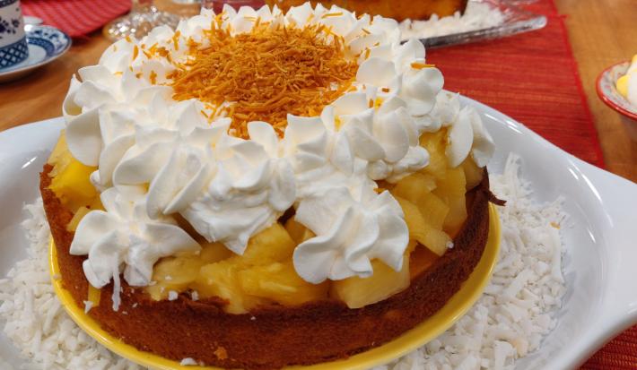 Foto aproximada da receita de bolo de pina colada com leite moça, um doce servido em um prato branco e amarelo, em camadas de massa, abacaxi picado, merengue decorado e coco ralado por cima.