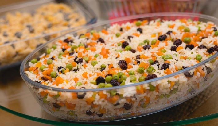 Em um recipiente de vidro transparente, há arroz branco misturado com cenouras, ervilhas e uvas-passas, decorado com duas folhas de hortelã.