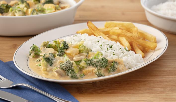 Fotografia em tons de verde em uma bancada de madeira com um guardanapo de pano azul, um prato oval branco no centro com o strogonoff de brócolis, arroz branco e batata frita. Ao fundo, um recipiente com strogonoff de brócolis e outro com arroz. Fotografia em tons de verde em uma bancada de madeira com um guardanapo de pano azul, um prato oval branco no centro com o strogonoff de brócolis, arroz branco e batata frita. Ao fundo, um recipiente com strogonoff de brócolis e outro com arroz.