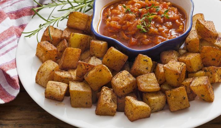 Fotografia em tons de vermelho em uma bancada de madeira escura com um pano branco com listras vermelhas e um prato branco raso grande ao centro com as batatas assadas e um potinho com o molho de tomate picante dentro.