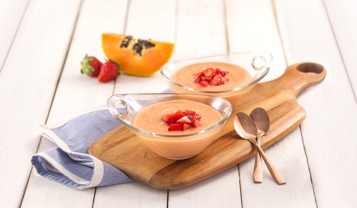 Fotografia em tons de azul e rosa em uma bancada de madeira branca, um paninho azul, uma tábua de madeira com duas taças em cima com creme de papaia dentro delas, decoradas com morangos. Ao fundo mamão e morangos.