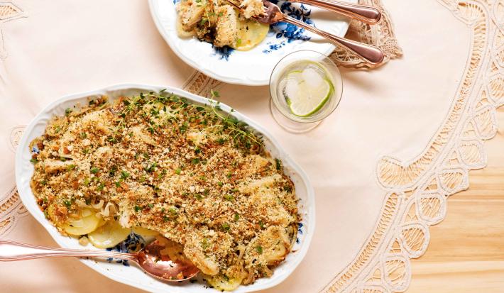 fotografia em tons de bege e branco de uma bancada vista de cima. Contém um pano branco, um recipiente oval branco com o bacalhau com uma colher para servir. Ao lado um prato redondo e branco com bacalhau e garfo e faca para servir