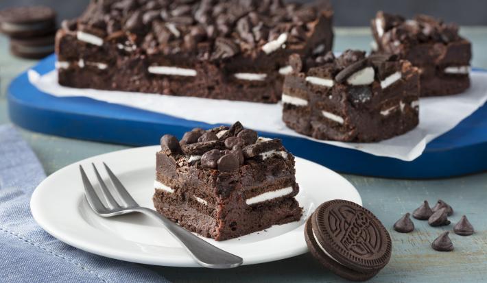 Fotografia em tons de azul em uma bancada de madeira cinza, um pano azul, uma tábua de madeira azul com um papel manteiga e o brownie com recheio de Biscoito Negresco dentro e em cima do papel. Ao centro, um prato branco redondo com um pedaço do brownie.