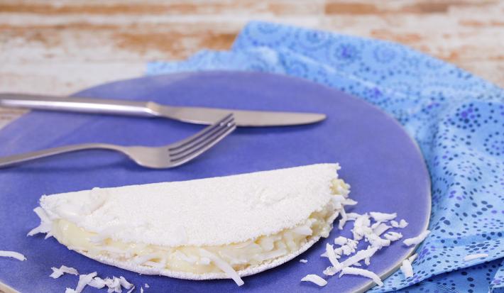 Fotografia em tons de azul em uma bancada de madeira clara, um pano azul, um prato azul redondo com uma tapioca de coco em cima dele.