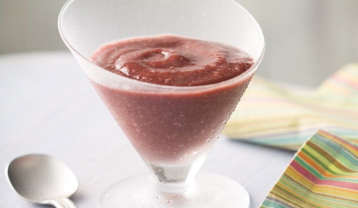 Foto de uma taça de vidro fosco com delícia de açaí de cor roxa intensa e textura cremosa, com leve redemoinho na superfície. A taça está sobre mesa clara, ao lado de uma colher prateada e de um guardanapo listrado colorido.