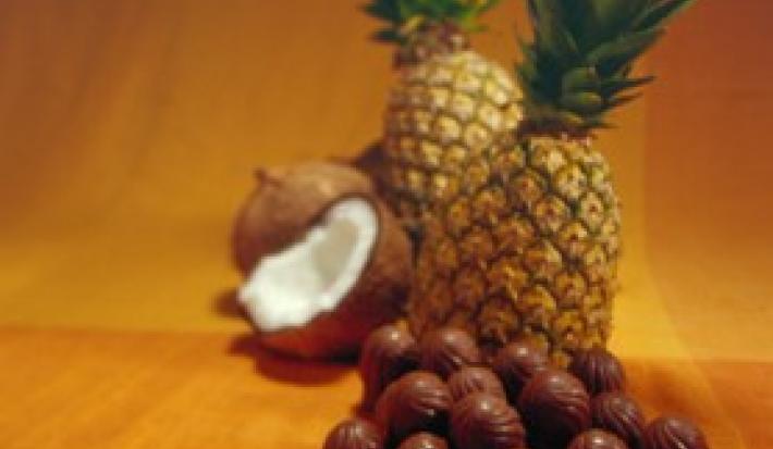 Fotografia em tons de marrom de uma bancada de marrom, sobre ela bombons, abacaxi e coco.