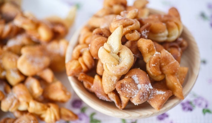 Em cima de uma mesa, recipiente redondo pequenos biscoitos em formato de laço.