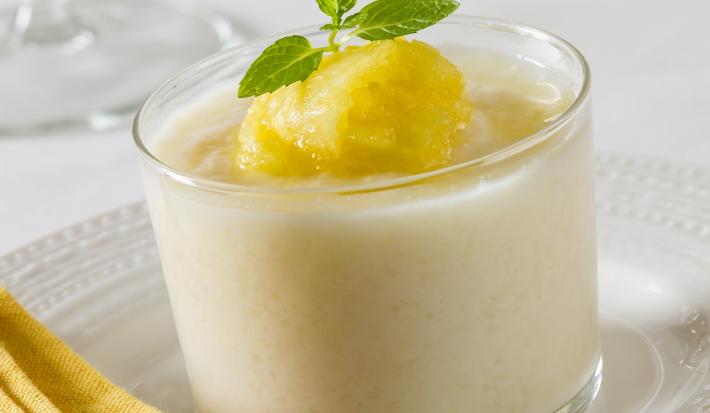 Fotografia de um recipiente pequeno transparente com um strogonoff doce de abacaxi gelado, com alguns pedaços sobre o creme. O recipiente está sobre um prato raso branco.