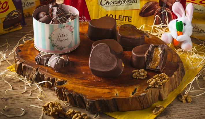 Foto de uma tábua de madeira escura com bombons de chocolate quadrados e em formatos de coração. Um deles está cortado ao meio mostrando o recheio. Tudo está em uma bancada decorada com um tecido amarelo, palha, um coelho de pelúcia e nozes espalhadas