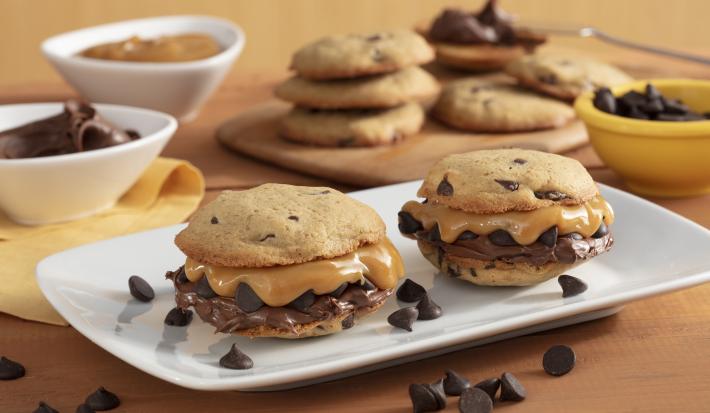 Fotografia em tons de amarelo em uma bancada de madeira com um recipiente branco ao centro com dois cookies recheados com doce de leite e creme de avelã. Ao fundo, potinhos com gotas de chocolate, doce de leite e uma tábua com mais cookies.