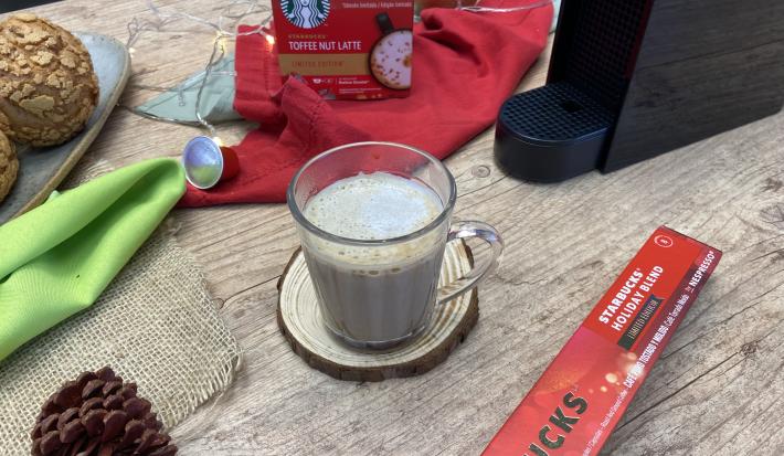 Fotografia em tons de vermelho em uma bancada de madeira com um pano vermelho, uma xícara de vidro com o café e enfeites natalinos.