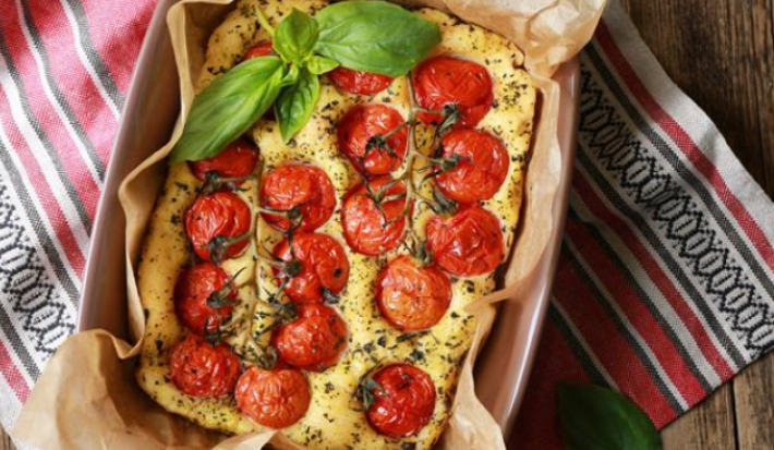 sobre um pano colorido listrado em tons de vermelho e branco está uma forma refratária com uma focaccia assada em tons dourados e toamtes cereja por cima. Folhas de manjericão guarnecem o prato.