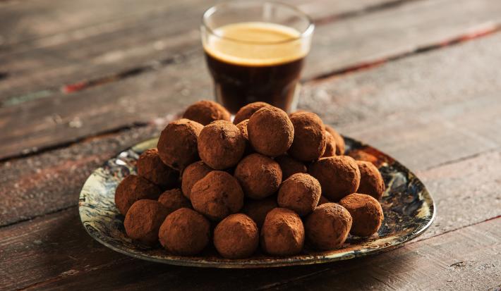 fotografia em tons de marrom e preto de uma bancada marrom vista de frente, contém um prato redondo com trufas com chocolate, e ao fundo um copo com café.