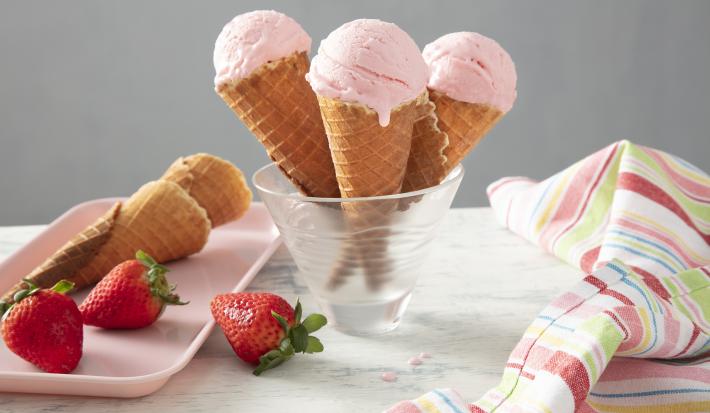 Foto da receita de Moça Sorvete de Nesquik. Observa-se um recipiente de vidro  com três casquinhas dentro, uma com cada bola. Ao lado esquerdo, casquinhas de sorvete e morangos decoram a foto sobre uma tábua rosinha.
