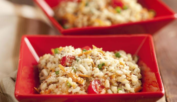 Fotografia em tons de vermelho, marrom e branco, com dois potinhos quadrados contendo um arroz colorido  e queijo ralado, ao lado guardanapo em tons de branco e marrom, tudo sobre bancada de madeira rústica.