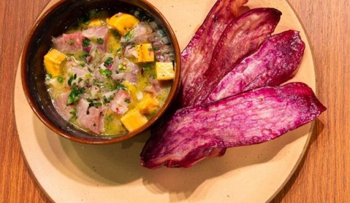 Fotografia vista de cima de um recipiente pequeno com ceviche ao lado de 4 lâminas de batata doce frita, sobre um prato de tom claro apoiado em uma bancada de madeira.