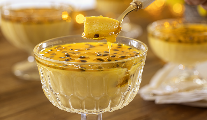 Fotografia apresenta uma mousse de maracujá servida em uma taça elegante. A mousse tem uma textura leve e cremosa, decorada com sementes de maracujá frescas e uma calda amarela