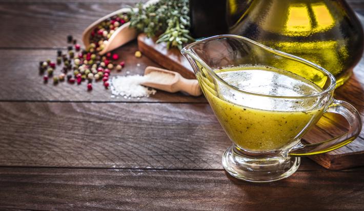Fotografia de um mini recipiente de vidro com um molho para vinagrete. Ao fundo, pimenta em grãos, sal e azeite, sobre uma mesa de madeira.