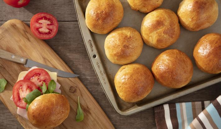 Imagem vista de cima de uma forma com vários pães redondos de batata-doce. Do lado esquerdo, um lanche montado com o pão, presunto, queijo, tomate e rúcula.