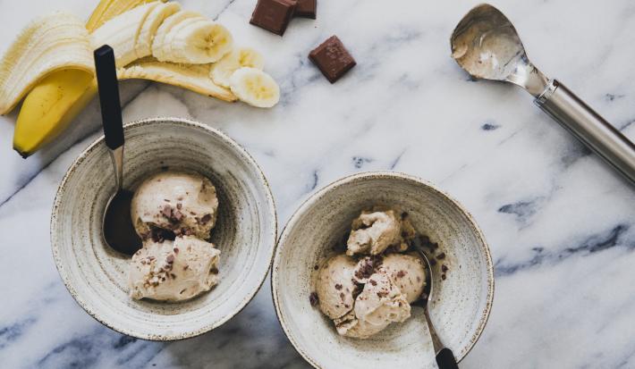 Fotografia em tons de amarelo, branco, azul e marrom de uma mesa branca e azul vista de cima, 2 recipientes redondos contém bolas de sorvete com pedaços de chocolates e cada um tem uma colher para servir. Ao fundo uma banana picada e pedaços de chocolate