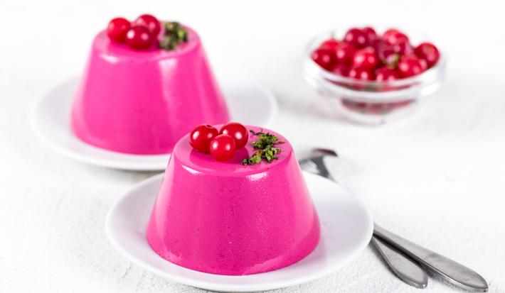 Fotografia de dois pratos pequenos com um manjar na cor rosa em cada, sobre eles, três cerejas para decoração. Ao fundo, várias cerejas em um refratário fundo de vidro pequeno, e ao lado dos pratos, duas colheres de sopa. Tudo está sobre uma mesa branca.