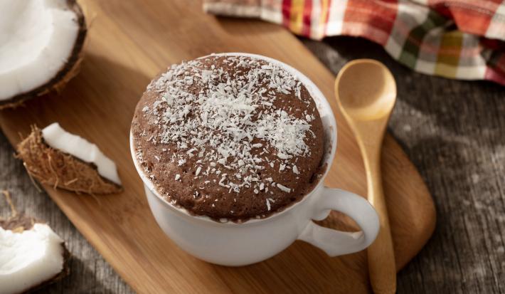 fotografia em tons de marrom e branco de uma bancada marrom vista de cima, contém uma tábua de madeira com uma xicara branca com bolo de prestígio e coco ralado por cima para decorar
