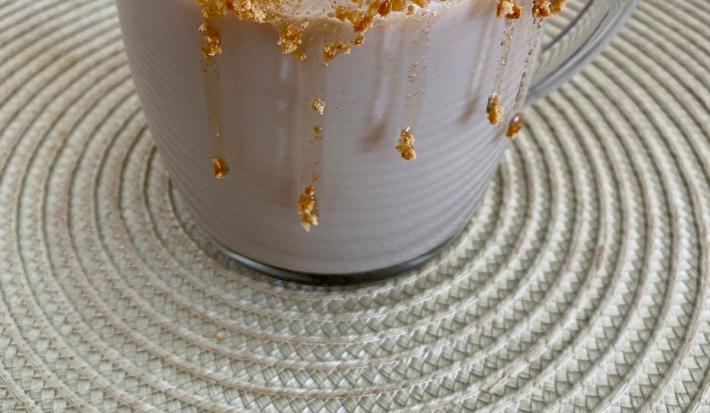 Fotografia em tons de marrom em uma bancada com um pano bege e uma xícara de vidro com a bebida de cappuccino de caramelo salgado dentro. Xícara decorada com calda de caramelo e biscoitos Tostines Especiarias