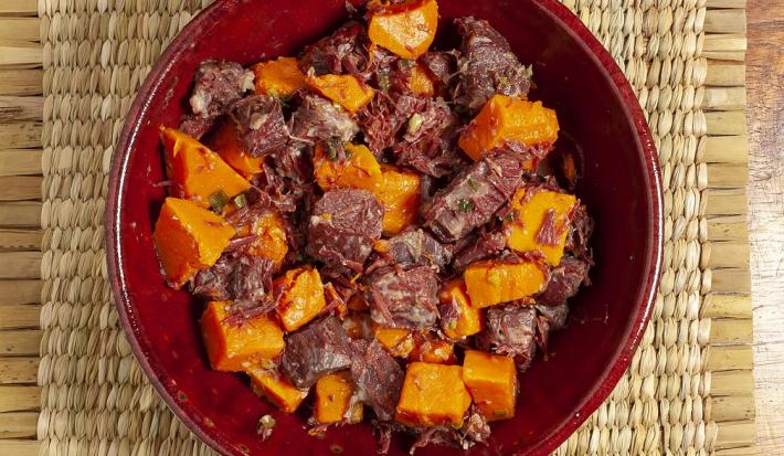 Fotografia em tons de amarelo, em uma bancada de madeira de cor marrom, ao centro um prato vermelho contendo a carne com abóbora.