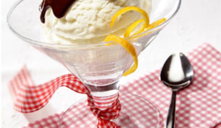 fotografia em tons de branco, vermelho e marrom de uma bancada branca vista de frente, contém um pano xadrez em tons de vermelho e branco. Uma taça transparente com uma bola de sorvete de creme com calda de chocolate