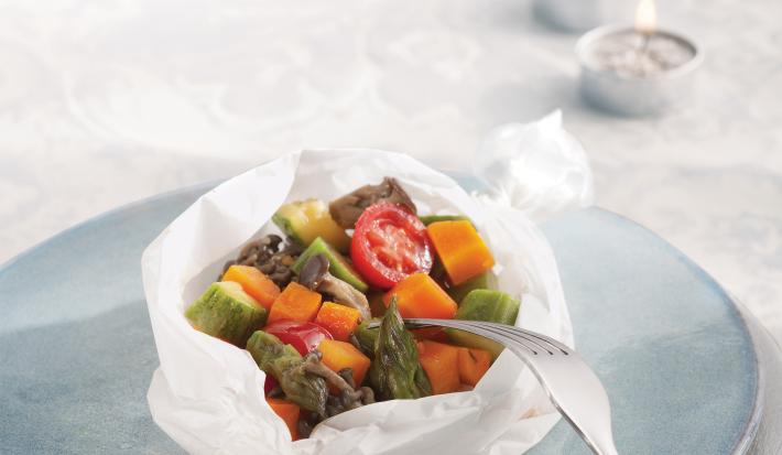 Fotografia em tons de azul em uma mesa com uma toalha branca de renda, um prato branco e azul claro em cima dela e o papillote de legumes (abobrinha, cenoura, tomate) em cima do prato azul, enrolado em um papel manteiga.