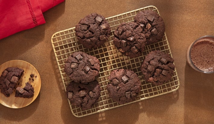 Fotografia de uma grelha quadriculada ao lado de um copo de vidro com leite e nescau e um mini apoio circular de madeira com um cookie ao meio. Sobre a grelha tem sete cookies de nescau extra cacau com gotas de chocolate. Fotografia de uma grelha quadriculada ao lado de um copo de vidro com leite e nescau e um mini apoio circular de madeira com um cookie ao meio. Sobre a grelha tem sete cookies de nescau extra cacau com gotas de chocolate.