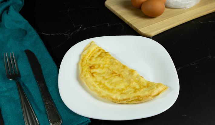 Fotografia em tons mais escuros de um prato branco com uma crepioca dobrada na metade. O prato está sobre uma mesa preta e ao lado de talheres, os quais estão apoiados em um pano de tom azul.