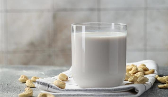 Fotografia em tons claros de um copo de vidro com uma bebida vegetal de castanha de caju apoiado em um pano branco dobrado com algumas listras escuras, que está sobre uma mesa de cimento queimado. Por cima da mesa e do pano tem castanhas espalhadas.
