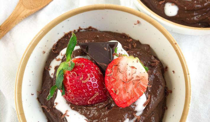 Fotografia em tons de vermelho em uma bancada de madeira com uma toalha branca, um prato branco fundo com detalhes dourados e o pudim de chocolate dentro dele, com raspas de chocolate e morangos para decorar.