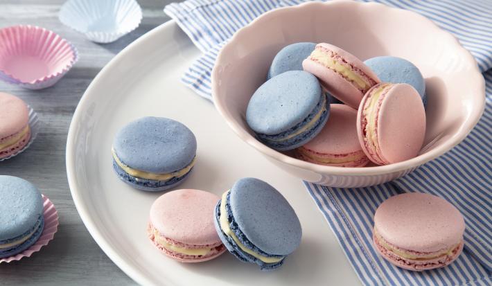 Imagem de macarons rosa e azul em um pote rosa claro sobre um prato branco. Ao lado mais macarons em cima do prato.